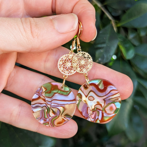 A pair of handmade dangle earrings held in a hand. The earrings feature gold filigree circular connectors and round drop pendants with a marbled, swirling pattern of purple, pink, green, and cream.