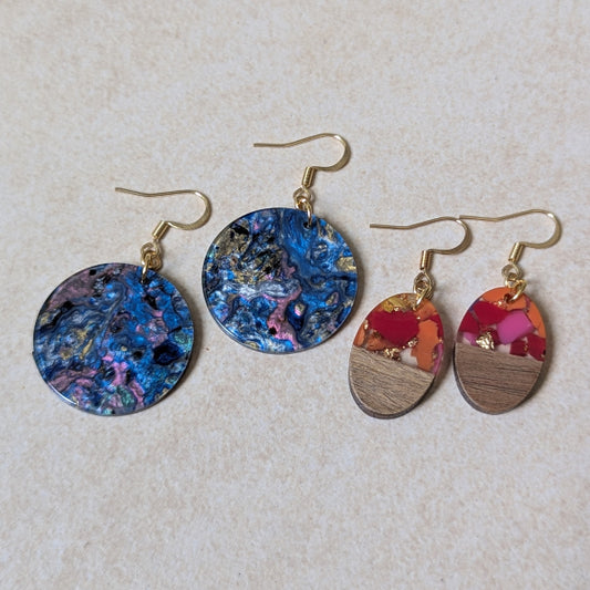 Two pairs of colourful earrings made from resin and wood on a beige background. Plated gold hooks.