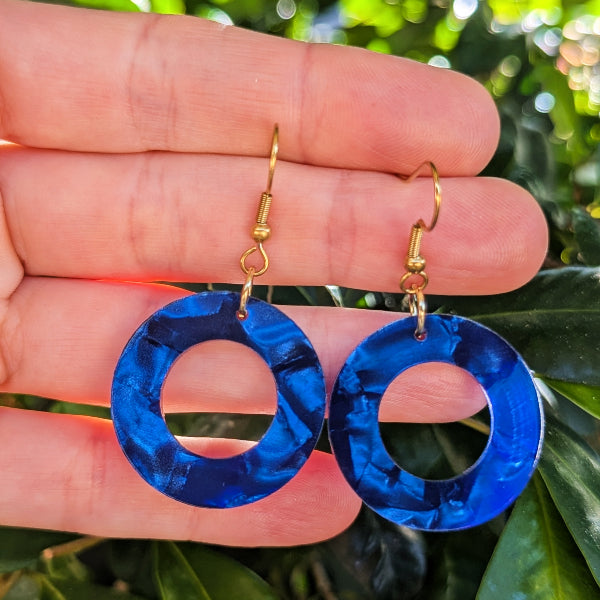 A pair of blue marbled circular drop earrings with gold hook wires, held in a person's hand against a background of green foliage.