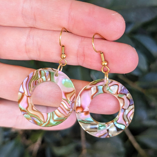 A pair of gold-tone dangle earrings with iridescent acrylic circular cutouts, held in a person's hand against a blurred green outdoor background.