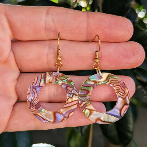 A pair of large circular hoop earrings with iridescent patterns and gold hook wires, held in a person's hand against a blurred outdoor background.