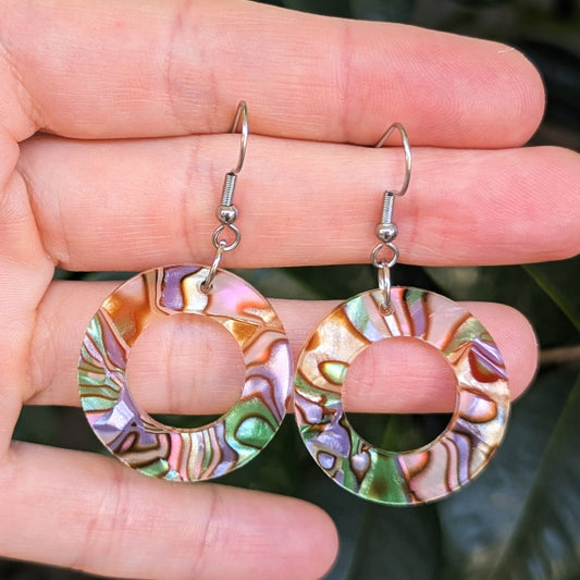 A pair of large circular hoop earrings with iridescent acrylic patterns in pink, green, gold, and purple, hanging from silver fishhook wires and held in a person's hand.