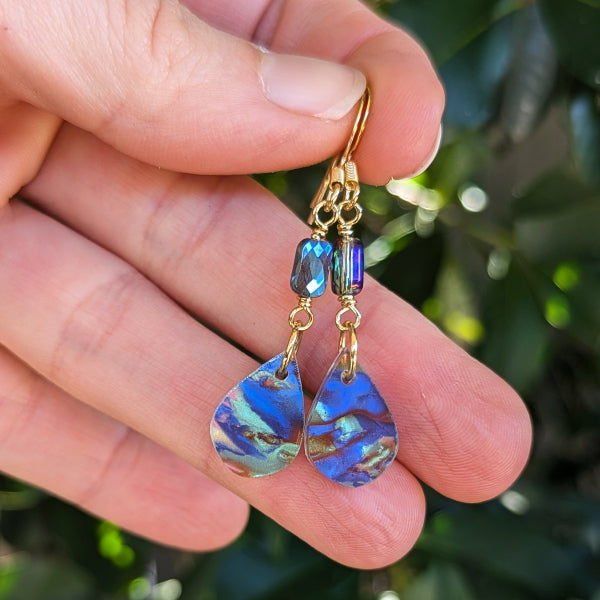 A pair of gold drop earrings with iridescent blue and green teardrop-shaped beads, held between fingers against a blurred green outdoor background.