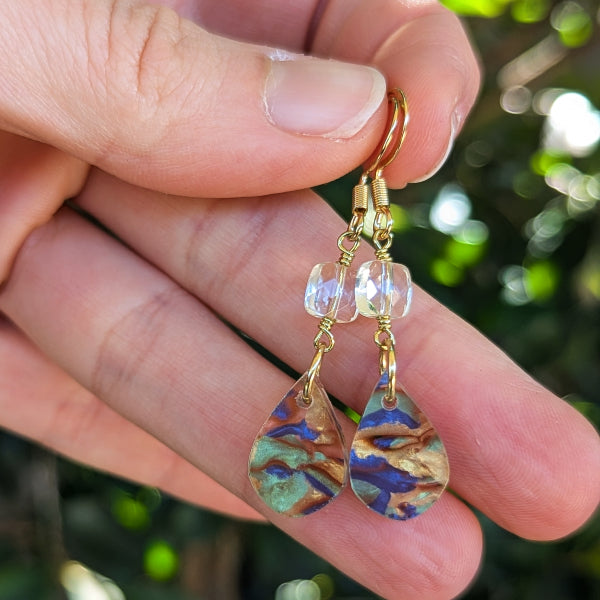 A pair of handmade teardrop dangle earrings held between fingers. The earrings feature gold-tone hook wires, clear faceted beads, and marbled acrylic drops with swirls of blue, green, and gold.