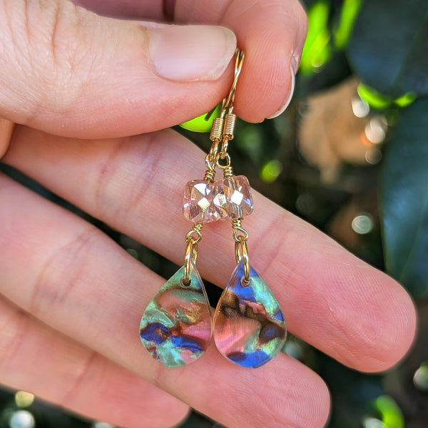A pair of gold drop earrings with faceted blush pink beads and teardrop-shaped iridescent charms, held in a person's hand against a blurred green outdoor background.