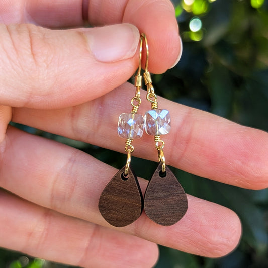 A pair of handmade teardrop dangle earrings held in a hand. The earrings feature pale blue faceted crystal beads and gold wire-wrapping with dark brown wooden teardrop pendants.