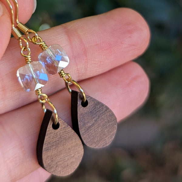 Wooden teardrop dangle earrings with faceted pale blue beads and gold-tone hooks, held in a hand against a blurred outdoor background.