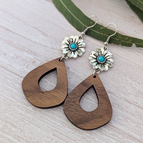 A pair of bohemian-style dangle earrings featuring dark brown wooden teardrop shapes with cutouts, connected to silver-toned flower charms with turquoise centers, displayed on a light wood surface with a green leaf.