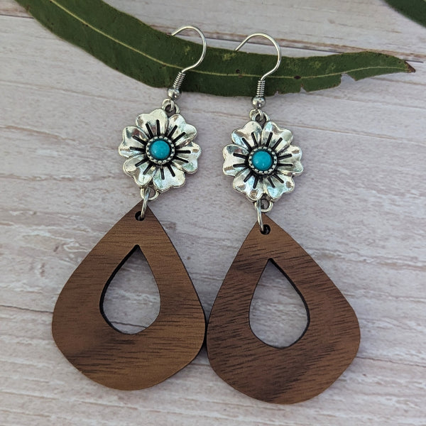 A pair of handmade dangle earrings featuring silver-toned flower charms with turquoise centers and large teardrop-shaped wooden drops, displayed on a light wood surface with a green leaf.