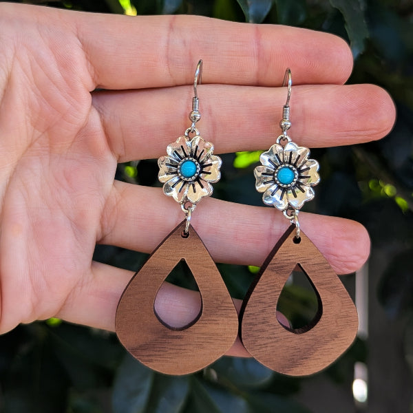 A pair of handmade dangle earrings held in a hand. The earrings feature silver-toned metal flower charms with turquoise blue stones, suspended from silver fishhook wires, and hang from large, open teardrop-shaped wooden pendants in a medium brown wood tone.