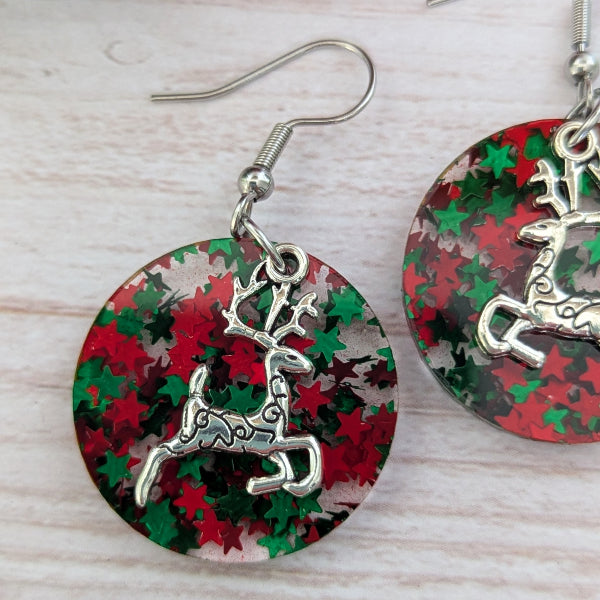 Round dangle earrings with a silver deer charm over a red and green star-patterned background, displayed on a light wood surface.