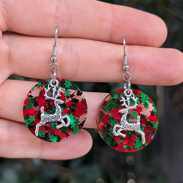 A pair of round dangling earrings held in a person's hand. Each earring has a silver-tone reindeer charm over a circular red and green glittery background.