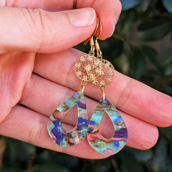 A close-up of a hand holding a pair of dangling earrings. The earrings have gold-tone hooks and filigree tops, with teardrop-shaped dangles featuring an iridescent acrylic pattern in blue, green, and gold.