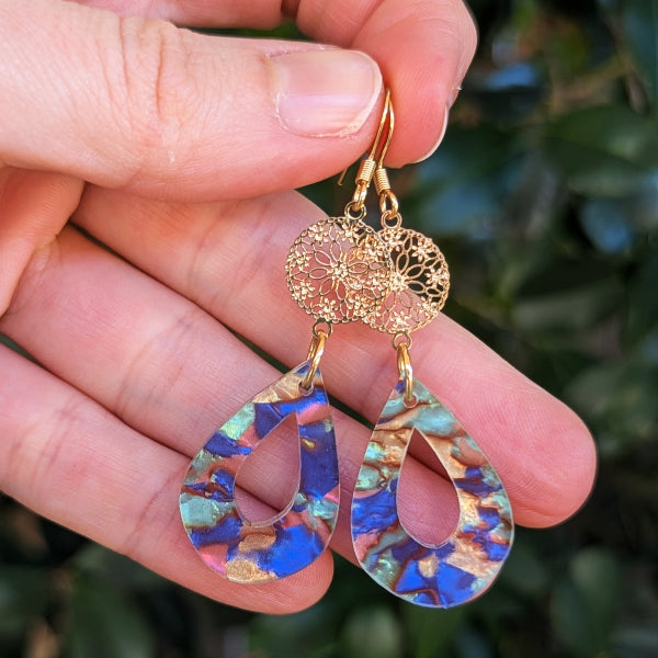 A pair of handmade teardrop dangle earrings held in a hand. The earrings feature a gold-tone filigree top and a marbled acrylic drop with blue, pink, and gold patterns.