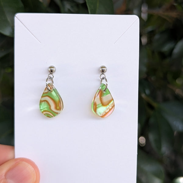 A pair of teardrop-shaped dangle earrings with marbled green and pink patterns, suspended from silver studs and held on a white card.