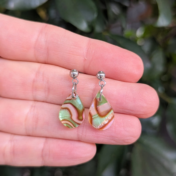 A pair of teardrop-shaped dangle earrings with marbled green and pink patterns, suspended from silver studs and held on a person's fingers against a blurred green background.
