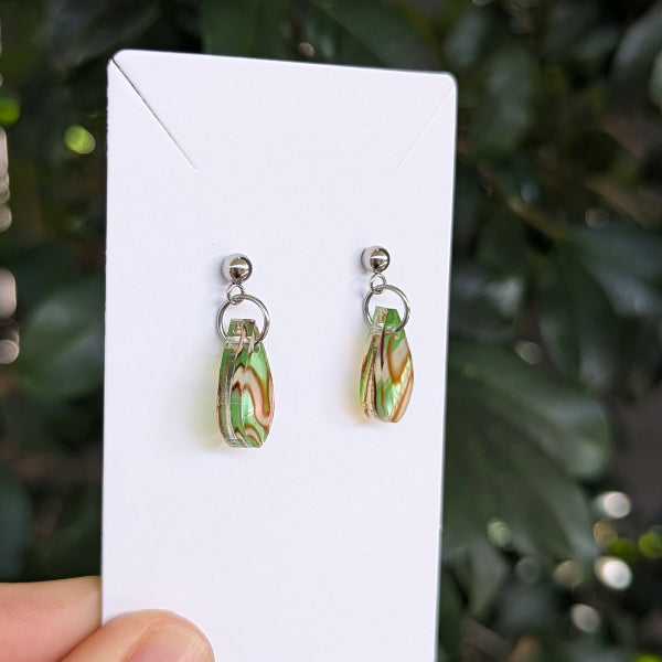 A pair of teardrop-shaped dangle earrings with marbled green and pink patterns, suspended from silver studs and held on a white card.