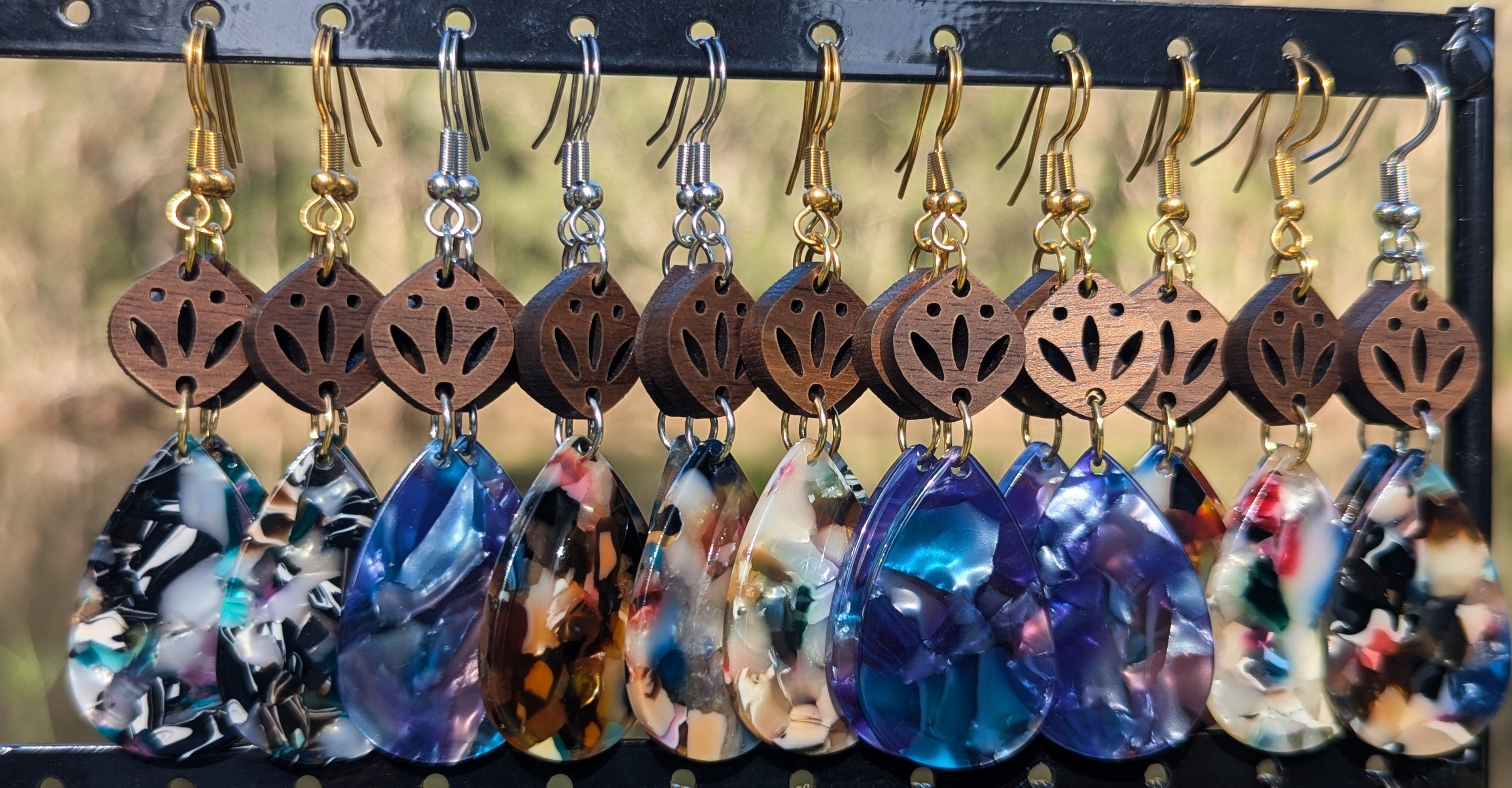 A row of handmade teardrop-shaped dangling earrings hanging from a black display rack. Each earring has a brown wooden top with a cutout floral pattern, a gold-tone or silver-tone hook, and a colorful marbled resin drop in shades of blue, purple, black, white, and orange.