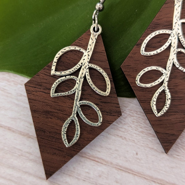 Wooden geometric dangle earrings with a silver leaf branch design, displayed on a light wood surface with a green leaf in the background.