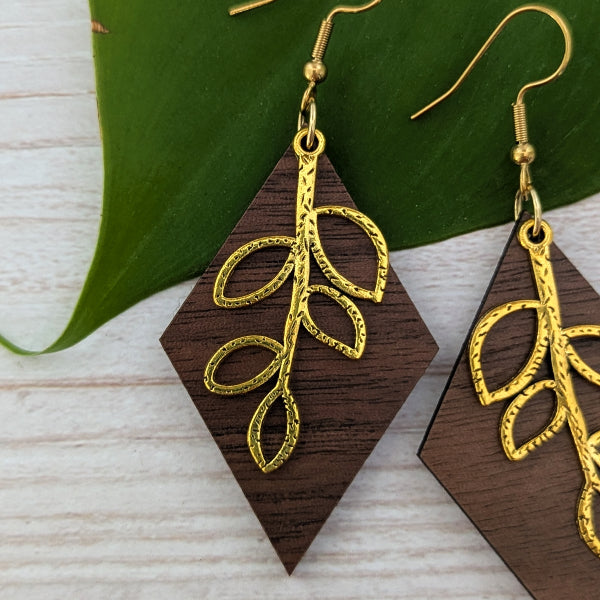 Wooden geometric dangle earrings with gold leaf filigree details, displayed on a light wood surface with a green leaf prop.
