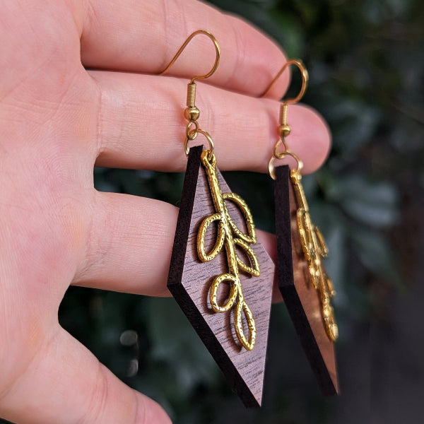 A close-up of a hand holding a pair of geometric wooden drop earrings with gold-tone hooks and leaf-shaped charms, set against a blurred green outdoor background.