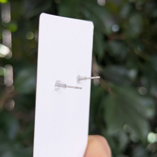 A close-up of a white display card holding two small clear rubber stud backs with silver posts, held by a hand against a blurred green outdoor background.