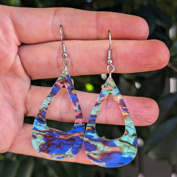 A pair of teardrop-shaped dangling earrings with a marbled blue, green, and copper pattern, held in a person's hand against a blurred green background.