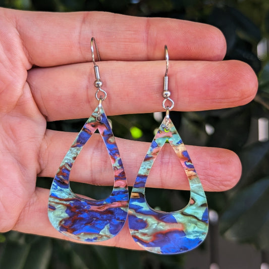 A pair of teardrop-shaped dangling earrings with a marbled blue, green, and copper pattern, held in a person's hand against a blurred green background.
