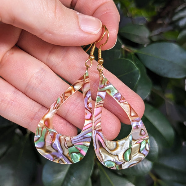A close-up of a hand holding a pair of teardrop-shaped earrings made from iridescent acrylic with gold hooks, set against a background of green leaves.