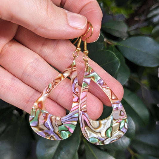 A close-up of a hand holding a pair of teardrop-shaped earrings made from iridescent acrylic with gold hooks, set against a background of green leaves.