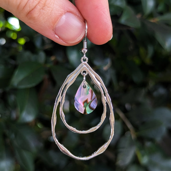 A hand holding a pair of silver dangle earrings with a teardrop-shaped iridescent acrylic pendant against a background of green foliage.