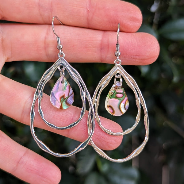 A pair of silver-toned double-ring teardrop dangle earrings with iridescent acrylic teardrop pendants, held in a person's hand against a blurred green outdoor background.