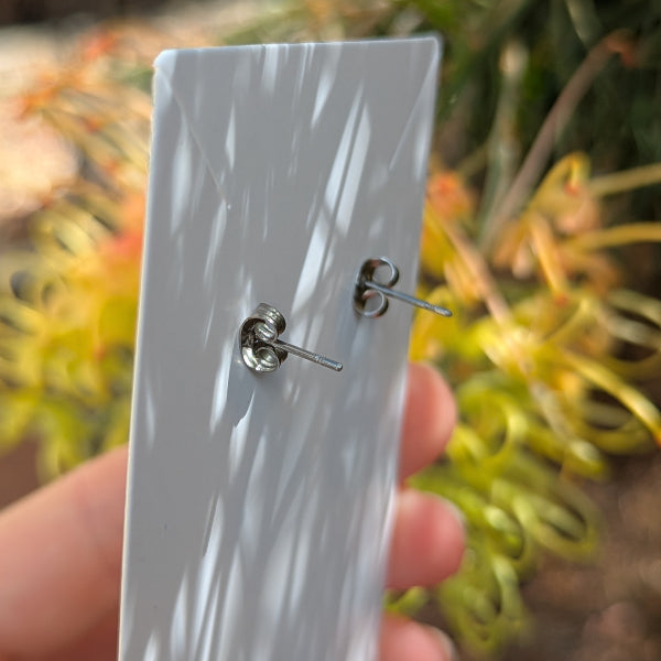 A pair of silver stud earrings displayed on a white rectangular card, held by a hand with a blurred outdoor garden background.