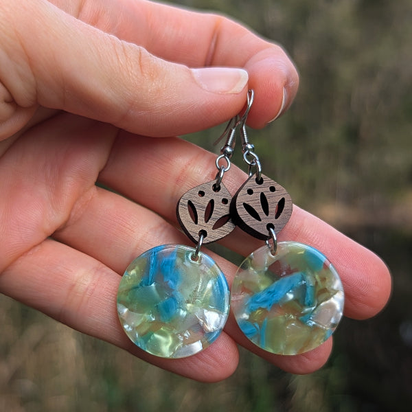 A hand holding a pair of handmade dangle earrings with round iridescent blue-green glass discs and dark brown wooden charms, against a blurred outdoor background.