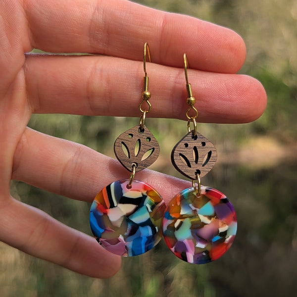 A close-up of a hand holding a pair of dangling earrings against a blurred outdoor background. The earrings have gold-tone fishhook wires, round cutout wooden discs, and colorful marbled circular drops in blue, red, orange, yellow, green, and purple.