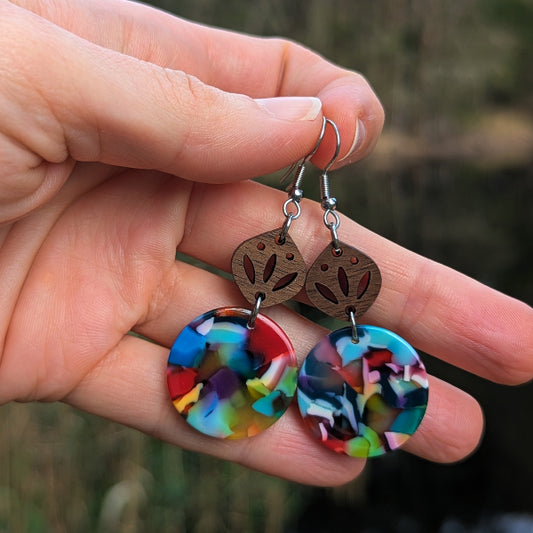 A close-up of a hand holding a pair of dangling earrings. The earrings have round, multicolored resin drop charms with a marbled pattern of red, blue, yellow, green, and black, suspended from small, laser-cut wooden discs with a leaf design, held by silver toned hook wires.