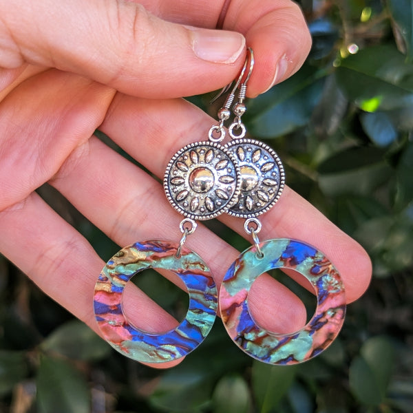 A pair of dangling earrings held in a hand against a blurred green outdoor background. The earrings have silver-toned sunburst charms and circular dangles with a marbled blue, green, pink, and gold pattern.