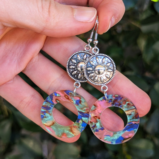 A pair of dangling earrings held in a hand against a blurred green outdoor background. The earrings have textured silver-tone sunburst charms and large circular dangles with a multicolor iridescent shell pattern showing blue, pink, green, and gold tones.