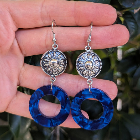 A pair of dangling earrings held in a hand, featuring round blue marbled hoop drops and silver-toned circular sunburst charms with a central flower detail, against a blurred background of green leaves.