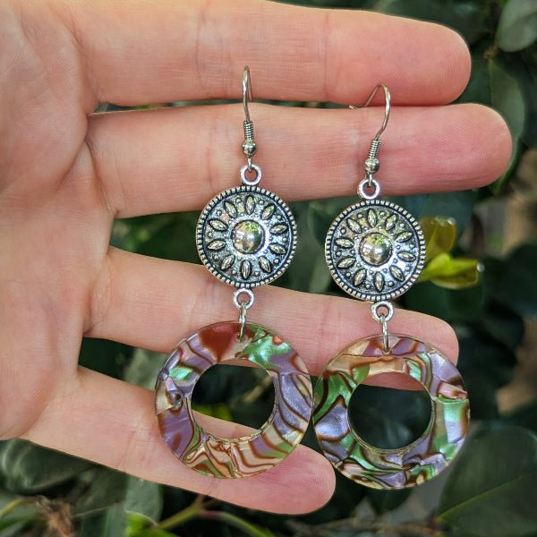 A pair of dangling earrings held in a hand against a blurred background of green leaves. The earrings feature silver-toned filigree round connectors with circle iridescent acrylic drops showing purple, green, and gold tones.