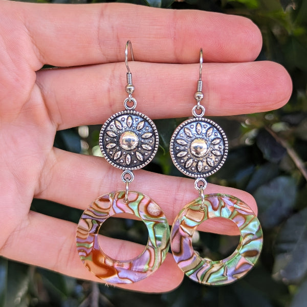 A pair of handmade dangle earrings held in a hand. The earrings feature textured silver-tone circular connectors with sunburst details and large, iridescent green, purple, and gold-toned circular drop charms.