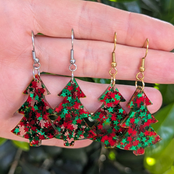 Four Christmas tree-shaped dangling earrings with red and green glitter, held in a person's hand against a blurred green outdoor background.