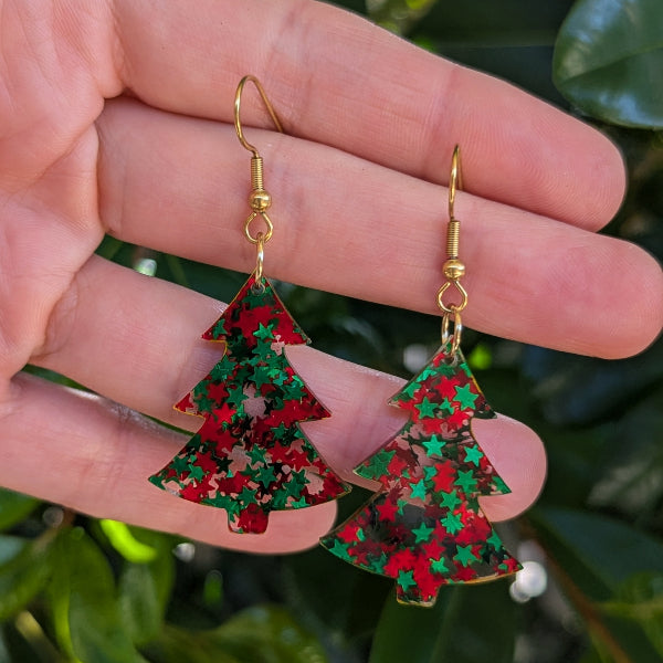 A pair of dangling earrings shaped like Christmas trees, decorated with red and green glitter and gold-tone hooks, held in a person's hand against a background of green leaves.
