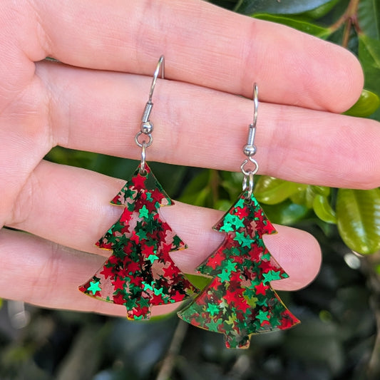 A pair of dangling earrings shaped like Christmas trees, covered in red and green glitter, held in a person's hand with blurred green leaves in the background.