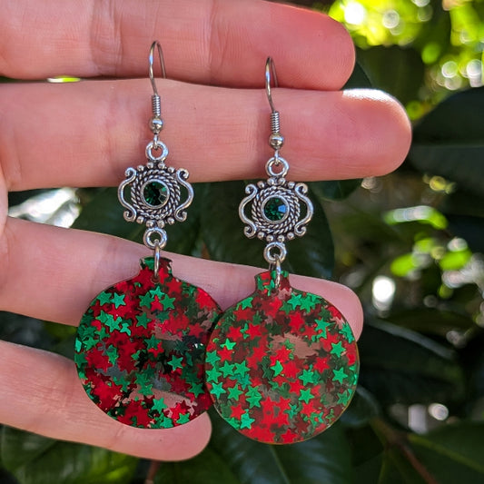 A pair of dangling earrings held in a hand, featuring round red and green star-patterned disc charms with ornate silver-toned hooks and small dark green stones.