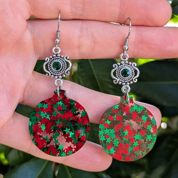 A pair of dangling earrings held in a hand, featuring round red and green star-patterned disc charms with ornate silver-toned hooks and small dark green stones.