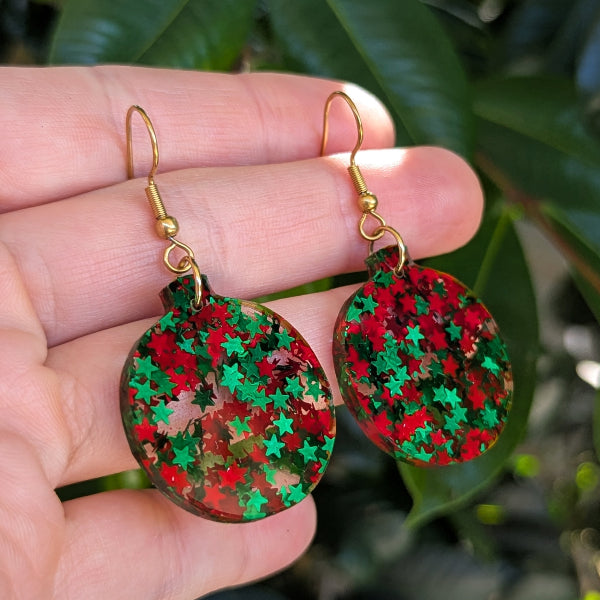 A pair of handmade dangle earrings with gold fishhook wires and round resin pendants covered in red and green star-shaped glitter, held in a person's hand against a background of green leaves.