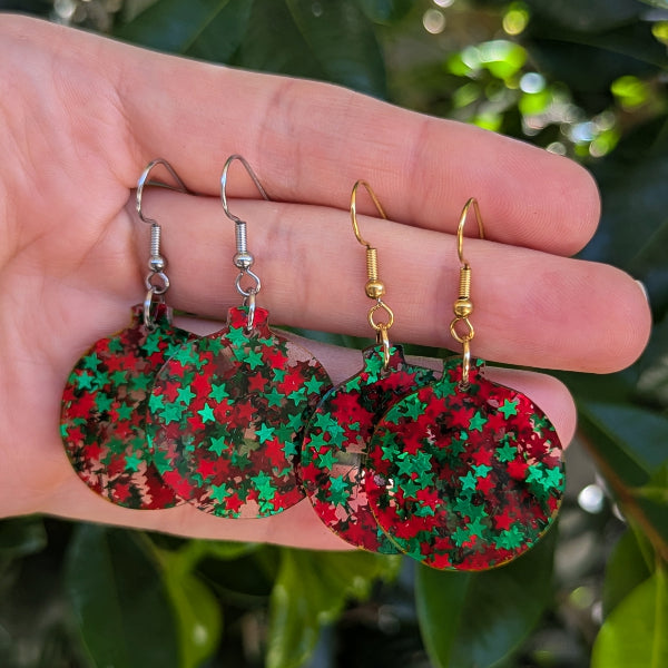 Two pairs of dangling round earrings with red and green star-patterned resin discs and silver and gold hooks, held in a person's hand with a blurred green leaf background.