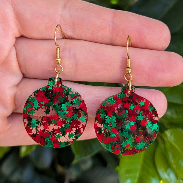 A pair of round dangling earrings with gold fishhook wires and a red and green star-patterned glitter finish, held in a person's hand against a blurred green leaf background.