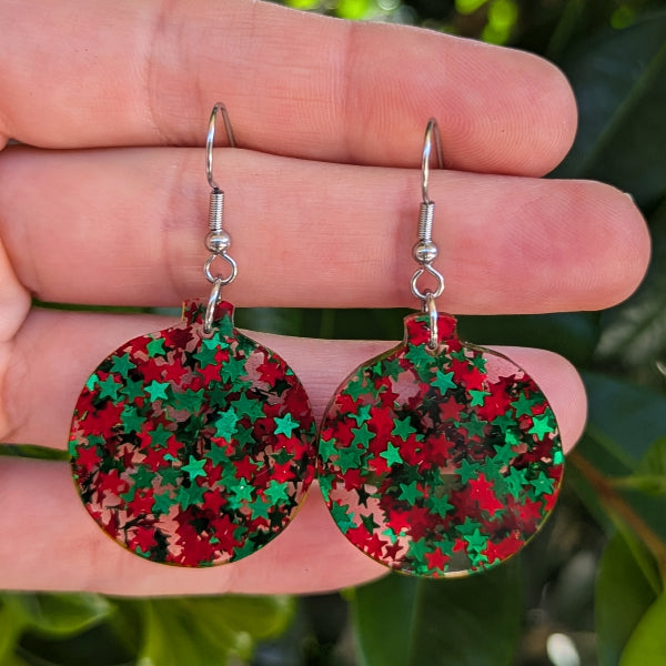 A pair of round silver-tone dangle earrings covered in red and green star-shaped glitter, held between a person's fingers against a blurred green outdoor background.
